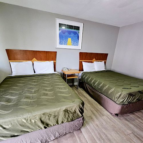 A hotel room with two double beds, green bedspreads, a bedside table, and a wall painting above the beds.