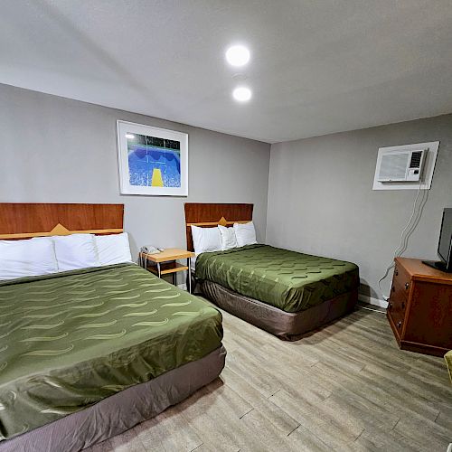 A hotel room with two double beds, green bedspreads, a nightstand, a wall-mounted AC unit, and a TV on a wooden stand.