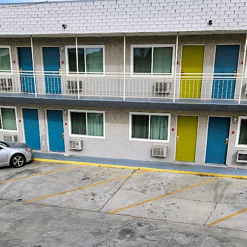 A two-story motel with colorful doors, a silver car, and a pickup truck parked in front, with stairs on both sides.