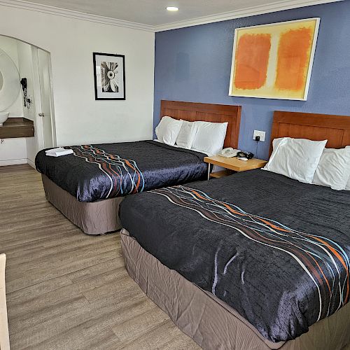 The image shows a hotel room with two double beds, modern decor, a circular mirror by the sink, and framed artwork above the beds.