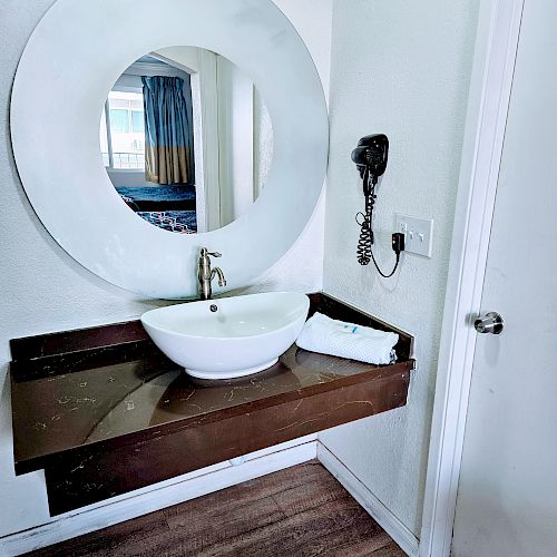 The image shows a bathroom vanity with a round mirror above, a white sink bowl, a hairdryer on the wall, and a towel on the wooden countertop.