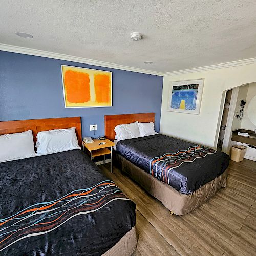 The image shows a hotel room with two double beds, modern decor, a nightstand in between, artwork on the walls, and wood flooring.