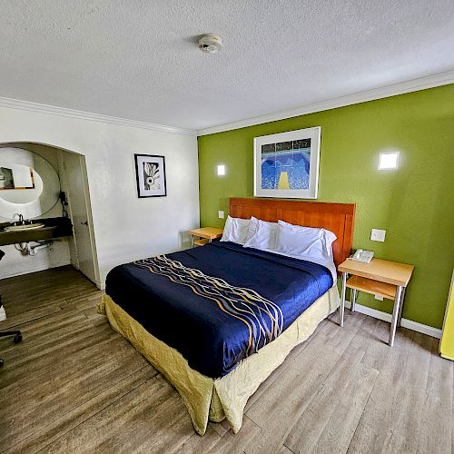 A hotel room with a queen bed, two side tables, a desk, a mirror, and a green accent wall adorned with artwork.
