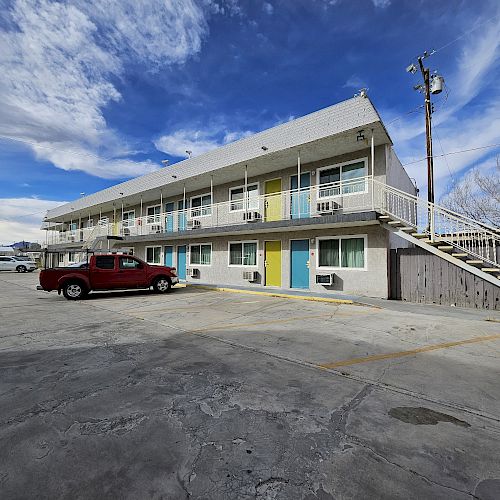 The image shows a two-story motel with colorful doors, a staircase, and parked vehicles under a partly cloudy sky in a spacious parking lot.