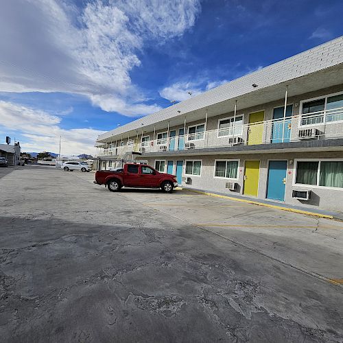 A two-story motel with colorful doors, a red truck, and several parked cars. The sky is partly cloudy with a strong blue color at places.