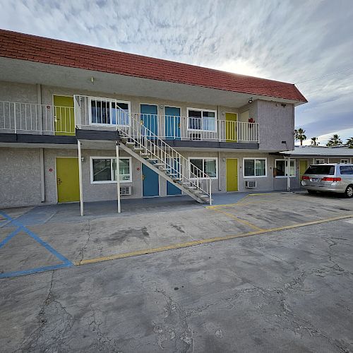 The image shows a two-story motel with colorful doors, an external staircase, a parking area, and a few parked cars under a cloudy sky.