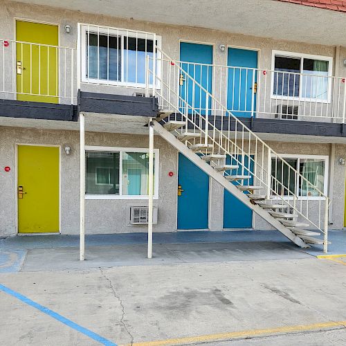 The image shows a two-story motel building with yellow and blue doors, a staircase in the center, and some parking spaces with blue lines on the ground.