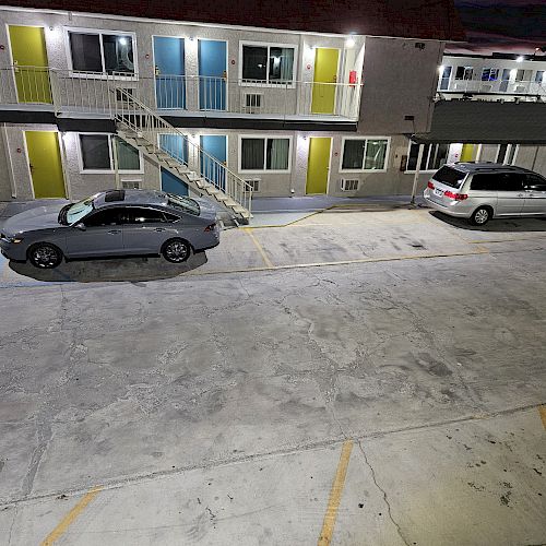 The image shows a parking area at night in front of a two-story motel with brightly colored doors and two cars parked in the lot.