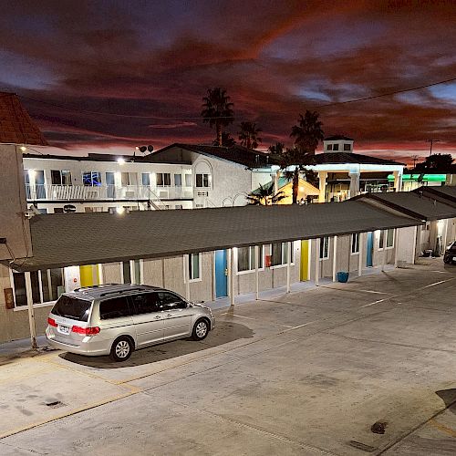 A row of motel rooms, two parked cars, and a dramatic evening sky with colorful clouds and distant buildings.