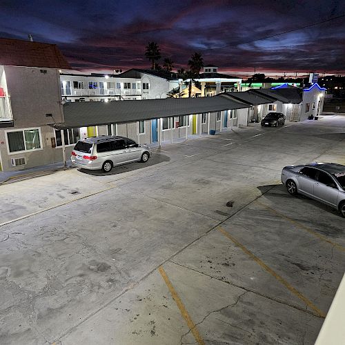 A motel parking lot at dusk with two cars parked; the sky is dark with some light from the building's windows and doors.