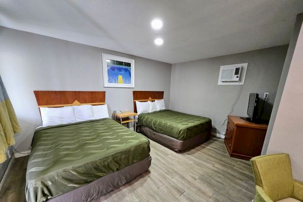 The image shows a hotel room with two double beds, a nightstand, wall art, an air conditioner, a TV on a wooden cabinet, and a green armchair.