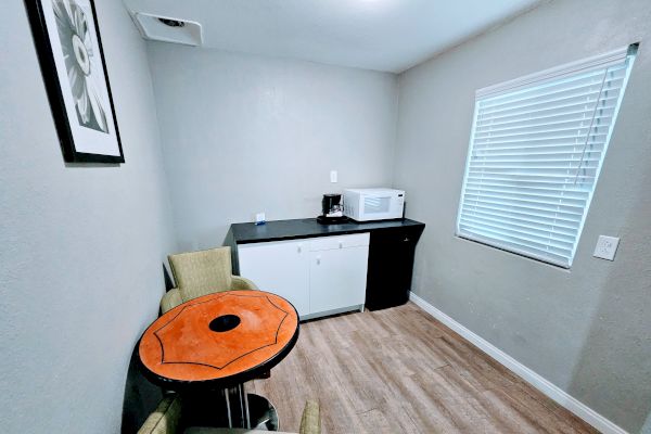 A small room with a table and two chairs, a kitchenette with a microwave and coffee maker, and a window with closed blinds.