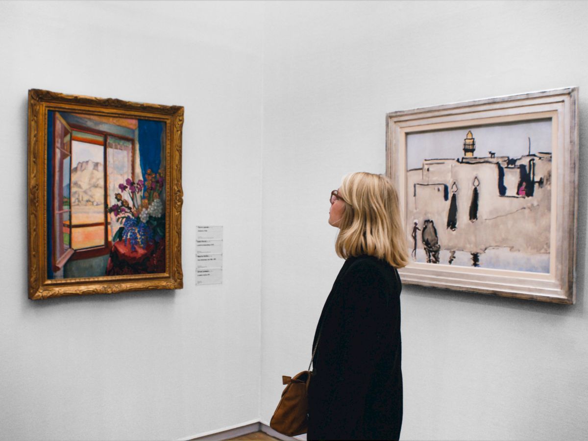 A person is observing two framed paintings on a gallery wall; one painting depicts a bouquet by a window and the other shows people in a city scene.